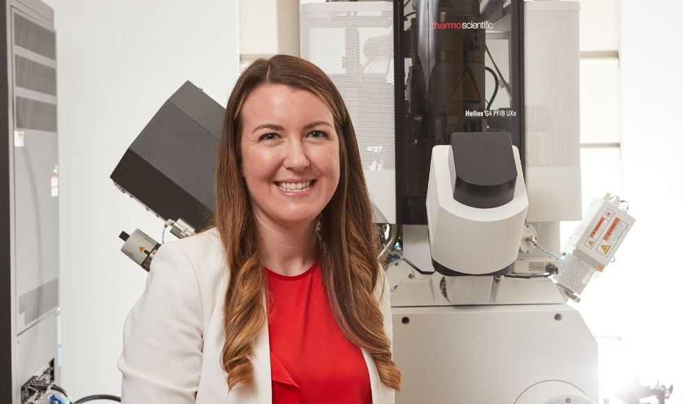 woman with long brown hair smiling in front of a microscope